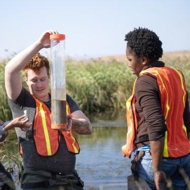 Two students are outside wearing reflective vests are gathering and observing water samples