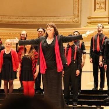 Dr Julie Ford, dressed in black-and-red, 当唱诗班站在她身后时，她向人群伸出双臂, arms to their sides