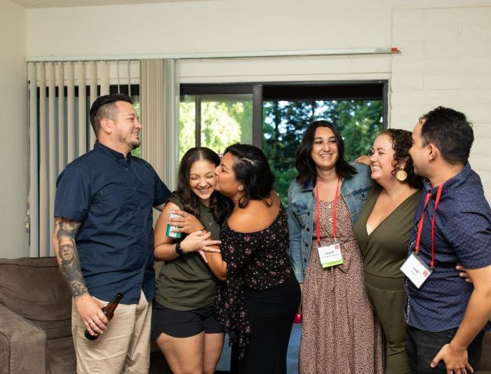 Six people embrace and laugh inside a townhouse living room